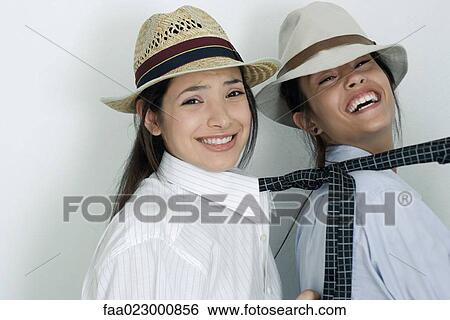 Stock Photograph - Two young female friends dressed in button down shirts, ties and hats, one pulling the other's tie, laughing, portrait. Fotosearch