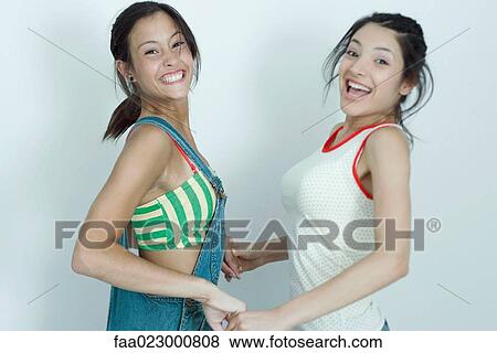 Stock Photo - Two young female friends holding hands and jumping, smiling at camera, portrait. Fotosearch