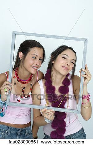Stock Image - Two young female friends holding up picture frame, smiling at camera, portrait. Fotosearch
