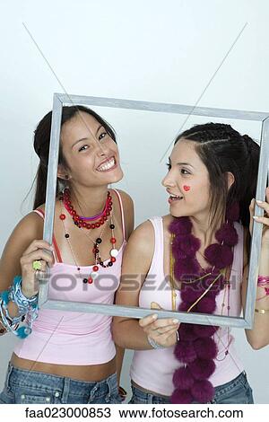 Stock Image - Two young female friends holding up picture frame, one making face at camera, portrait. Fotosearch