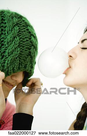 Two young female friends, one blowing bubble with chewing gum while the other pulls hat over eyes View Large Photo Image Stock Photo - Two young female friends, one blowing bubble with chewing gum while the other pulls hat over eyes. Fotosearch