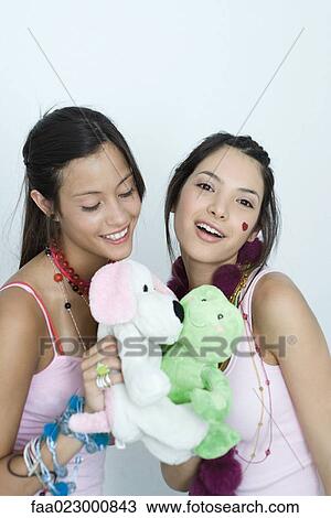 Stock Image - Two young female friends playing with stuffed animals, one smiling at camera, portrait. Fotosearch