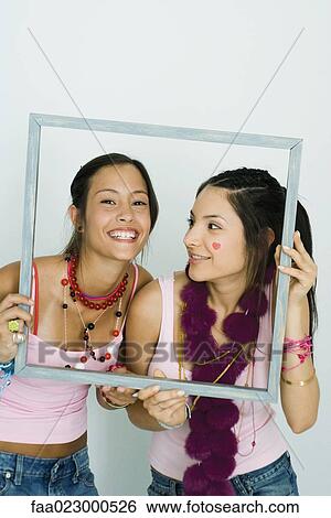 Stock Photograph - Two young friends holding up frame together, one smiling at camera, portrait. Fotosearch