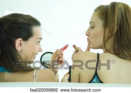 Stock Image - Two young friends holding up lipstick, one looking in hand mirror, rear view. Fotosearch