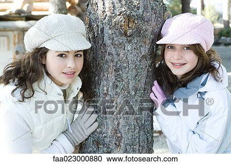 Stock Image - Two young friends leaning against tree trunk, both wearing caps, smiling at camera, portrait. Fotosearch