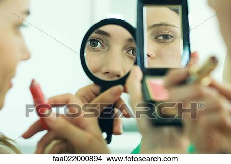 Picture - Two young friends looking at selves in hand mirrors, holding lipstick, cropped view. Fotosearch