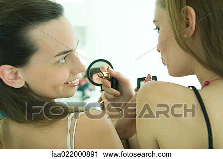 Stock Image - Two young friends putting on makeup, looking at each other, rear view. Fotosearch