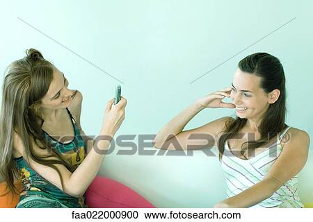 Two young friends sitting on couch, one taking photo of the other with cell phone View Large Photo Image Stock Image - Two young friends sitting on couch, one taking photo of the other with cell phone. Fotosearch