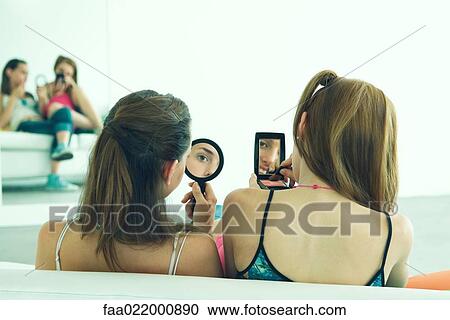 Stock Image - Two young friends sitting side by side, looking in hand mirrors, rear view. Fotosearch