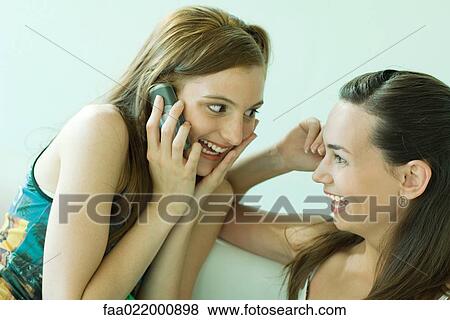 Two young friends smiling at each other, one using cell phone View Large Photo Image Stock Photo - Two young friends smiling at each other, one using cell phone. Fotosearch