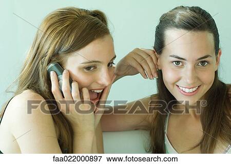 Stock Photo - Two young friends smiling together, using cell phone, one looking at camera. Fotosearch