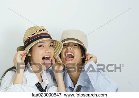 Stock Photo - Two young friends wearing hats, sticking out tongues, looking at camera, portrait. Fotosearch
