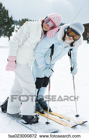 Stock Image - Two young skiers standing together, smiling at camera, one leaning on the other's shoulder, portrait. Fotosearch