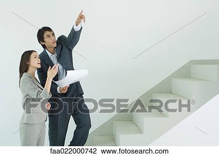 Stock Image - Well-dressed couple visiting house, man holding blueprint and pointing . Fotosearch