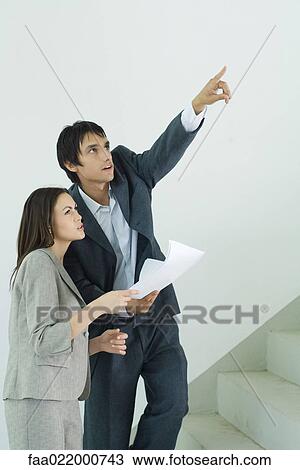 Stock Image - Well-dressed couple visiting house, man holding blueprint and pointing . Fotosearch