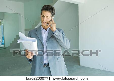 Stock Image - Well-dressed man looking at blueprints, using phone. Fotosearch