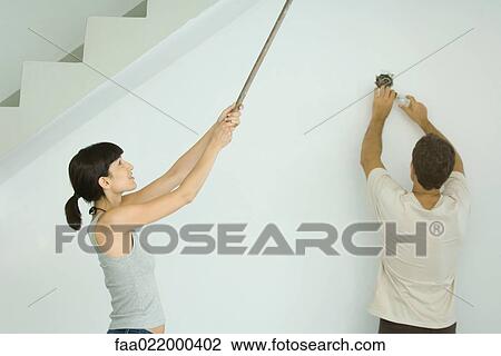 Stock Image - Woman painting ceiling with roller extension, man changing light bulb. Fotosearch