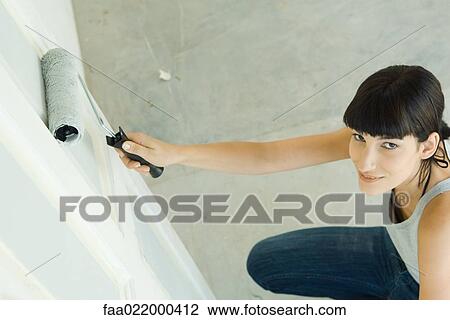 Stock Image - Woman painting door with paint roller, smiling up at camera. Fotosearch