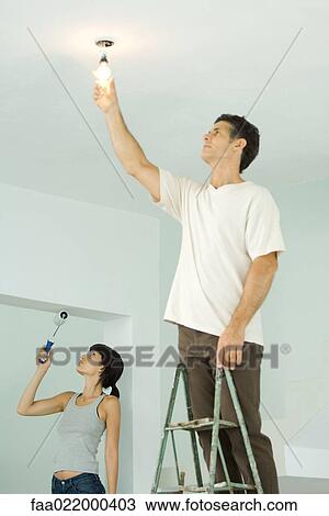 Stock Image - Woman painting with paint roller, man changing light bulb. Fotosearch