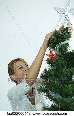 Stock Image - Woman placing star on top of Christmas tree. Fotosearch
