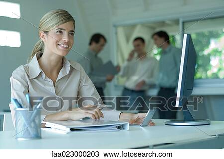 Woman sitting at desk, smiling, male colleagues standing in background View Large Photo Image Stock Image - Woman sitting at desk, smiling, male colleagues standing in background. Fotosearch