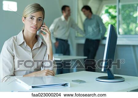Woman sitting at desk, using cell phone, male colleagues standing in background View Large Photo Image Picture - Woman sitting at desk, using cell phone, male colleagues standing in background. Fotosearch
