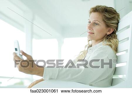 Stock Image - Woman sitting on bench, looking at cell phone, side view. Fotosearch