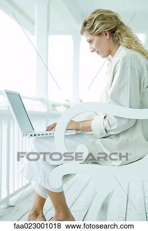 Stock Photo - Woman sitting on porch, using laptop computer, side view. Fotosearch