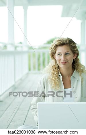 Stock Photo - Woman sitting on porch, using laptop computer, looking away, smiling. Fotosearch