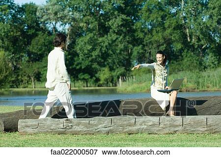 Stock Photo - Woman sitting on tree trunk, using laptop, motioning to approaching man. Fotosearch