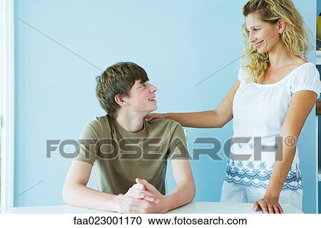 Woman standing next to teen son sitting at table, hand on his shoulder, both smiling at each other View Large Photo Image Stock Image - Woman standing next to teen son sitting at table, hand on his shoulder, both smiling at each other. Fotosearch