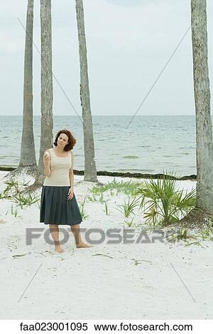 Stock Photography - Woman standing on the beach looking at cell phone, head tilted. Fotosearch