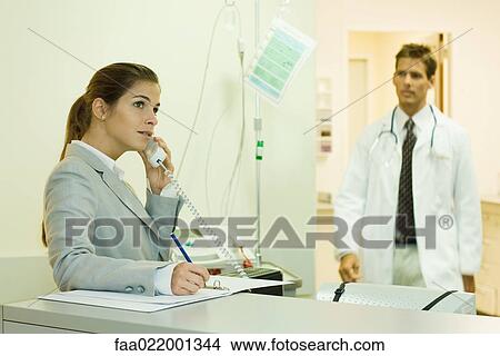 Picture - Woman using phone in doctor's office, doctor approaching in background. Fotosearch
