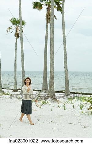 Stock Image - Woman walking on beach, looking down at cell phone, full length. Fotosearch