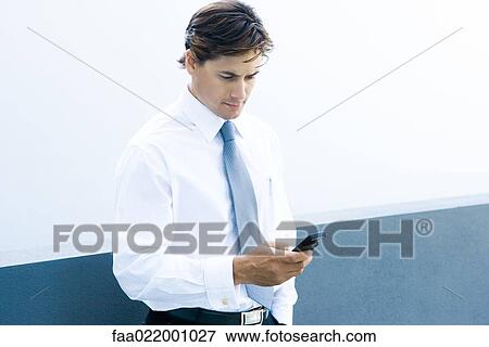 Stock Photo - Young businessman looking at cell phone, waist up. Fotosearch