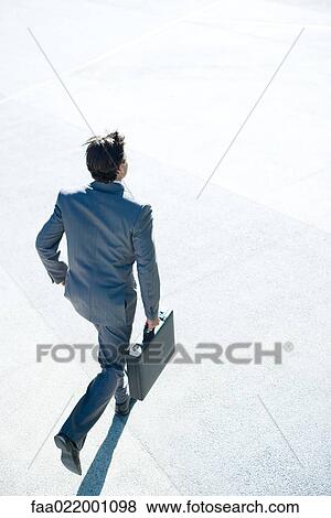 Stock Photo - Young businessman running outdoors, carrying briefcase, rear view, high angle view. Fotosearch