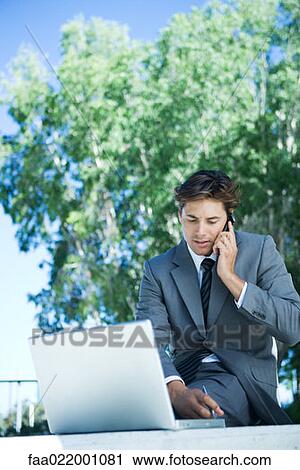 Young businessman sitting outdoors using cell phone and laptop computer, writing View Large Photo Image Stock Image - Young businessman sitting outdoors using cell phone and laptop computer, writing. Fotosearch