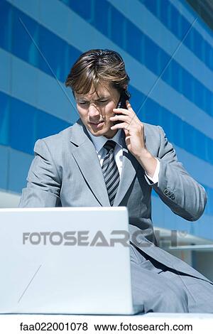 Young businessman sitting outdoors, using laptop computer, holding up cell phone View Large Photo Image Stock Photo - Young businessman sitting outdoors, using laptop computer, holding up cell phone. Fotosearch