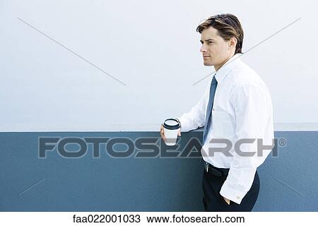 Stock Image - Young businessman standing, holding hot beverage, hand in pocket. Fotosearch