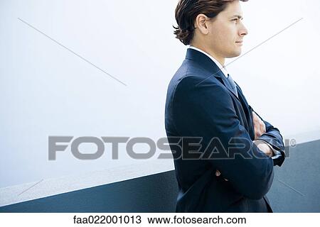 Stock Image - Young businessman standing with arms folded, cropped side view. Fotosearch