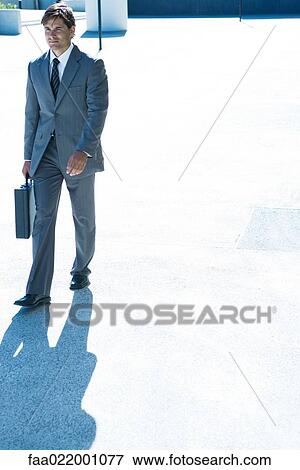 Stock Photo - Young businessman walking outdoors, carrying briefcase, full length. Fotosearch