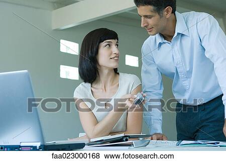 Young businesswoman sitting at desk, holding cell phone, glancing up at male colleague View Large Photo Image Stock Photo - Young businesswoman sitting at desk, holding cell phone, glancing up at male colleague. Fotosearch