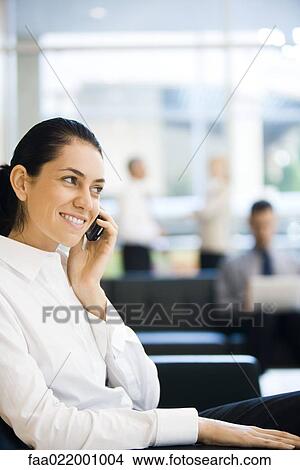 Young businesswoman sitting in lobby, using cell phone, smiling View Large Photo Image Picture - Young businesswoman sitting in lobby, using cell phone, smiling. Fotosearch
