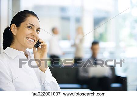 Stock Photo - Young businesswoman sitting in lobby, using cell phone, smiling, looking up. Fotosearch