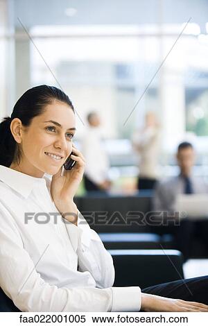 Stock Photography - Young businesswoman sitting in lobby, using cell phone, smiling. Fotosearch