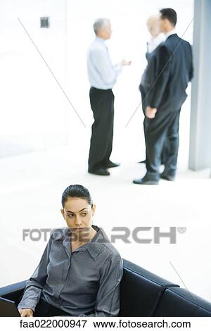 Young businesswoman sitting on sofa, looking away, associates in background View Large Photo Image Stock Photo - Young businesswoman sitting on sofa, looking away, associates in background. Fotosearch