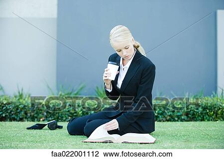 Stock Image - Young businesswoman sitting on the ground outdoors, reading book, holding hot beverage. Fotosearch