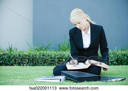 Stock Image - Young businesswoman sitting on the ground outdoors, reading book, holding pen. Fotosearch