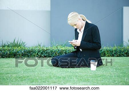 Young businesswoman sitting on the ground outdoors, looking at cell phone, hot beverage View Large Photo Image Stock Photo - Young businesswoman sitting on the ground outdoors, looking at cell phone, hot beverage. Fotosearch
