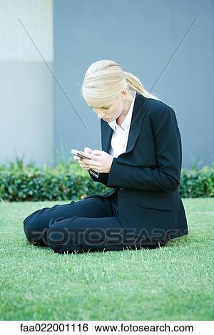 Young businesswoman sitting on the ground outdoors, looking at cell phone View Large Photo Image Stock Photograph - Young businesswoman sitting on the ground outdoors, looking at cell phone. Fotosearch